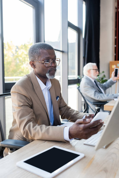 hombre de negocios afroamericano senior en anteojos mirando el teléfono inteligente mientras trabaja cerca de colega borrosa - Foto, imagen