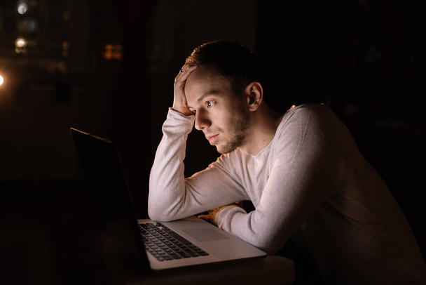 Młody człowiek wygląda na zmęczonego podczas pracy na laptopie w ciemnym biurze późnym wieczorem - Zdjęcie, obraz
