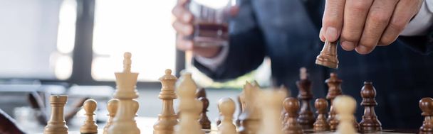 cropped view of businessman with blurred glass of whiskey playing chess in office, banner - Foto, Bild