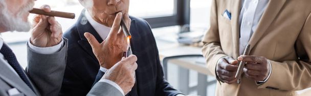 cropped view of senior businessman lighting cigar of business partner near blurred african american colleague, banner - Fotó, kép