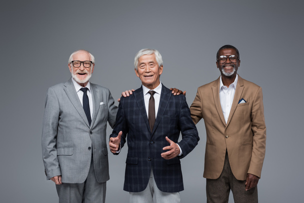 glimlachende multi-etnische zakenlieden aanraken schouders van senior aziatische collega geïsoleerd op grijs - Foto, afbeelding