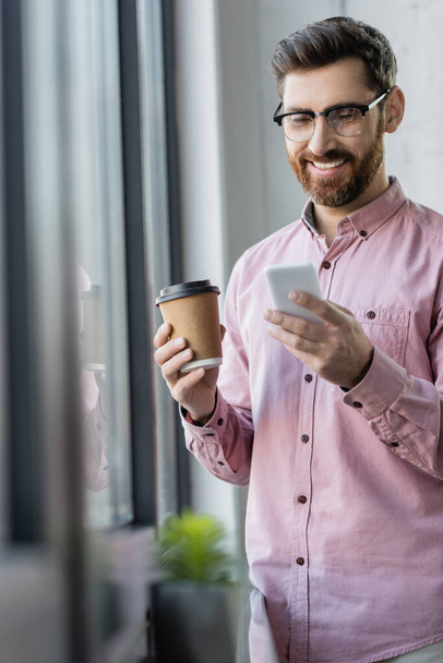 Uomo d'affari sorridente in possesso di caffè per andare e utilizzando smartphone in ufficio  - Foto, immagini