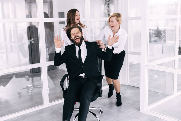Happy carefree women enjoy office activities, leading man - rolling in a chair, celebrating business success, great relationships, having fun during a break from work, laughing - Foto, Imagen