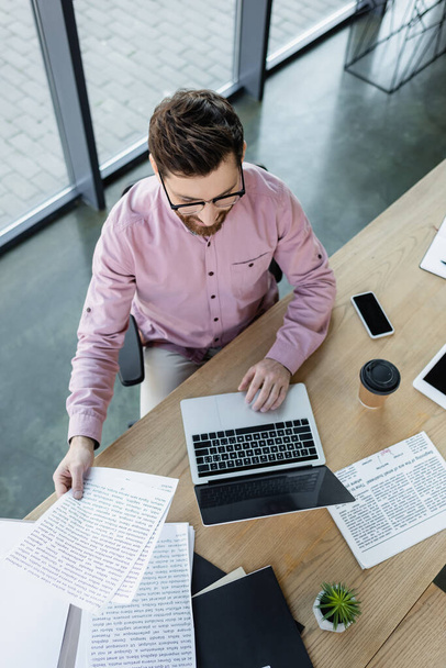 Overhead-Ansicht von Geschäftsmann mit Laptop und Arbeiten mit Papieren in der Nähe von Kaffee im Büro  - Foto, Bild