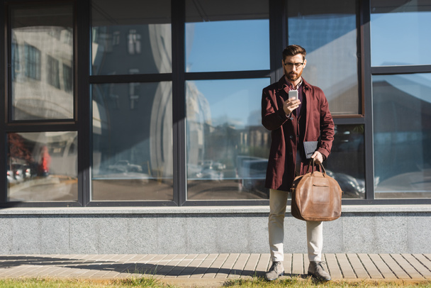Homme d'affaires dans un sac de maintien de trench coat et en utilisant un smartphone près du bâtiment à l'extérieur  - Photo, image