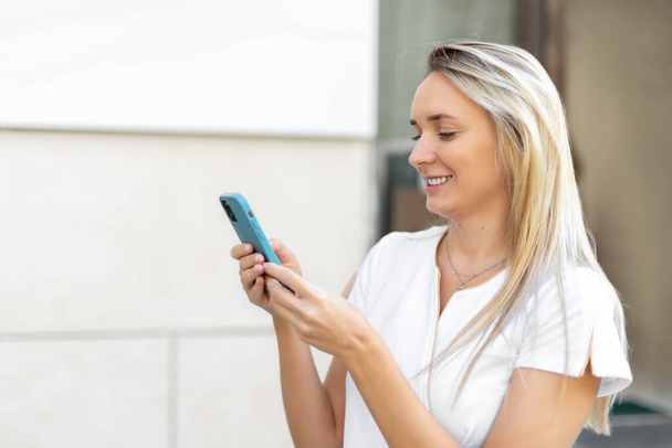 Κοντινό πλάνο πορτρέτο του νεαρού ενήλικα ευτυχισμένη γυναίκα πρόσωπο χρησιμοποιώντας smartphone στο δρόμο της πόλης σε εξωτερικούς χώρους για ηλιόλουστο πρωί. Όμορφη χαμογελαστή γυναίκα κρατάει το τηλέφωνο και βγαίνει έξω. Ξανθιά κοπέλα που κοιτάει στην άκρη - Φωτογραφία, εικόνα