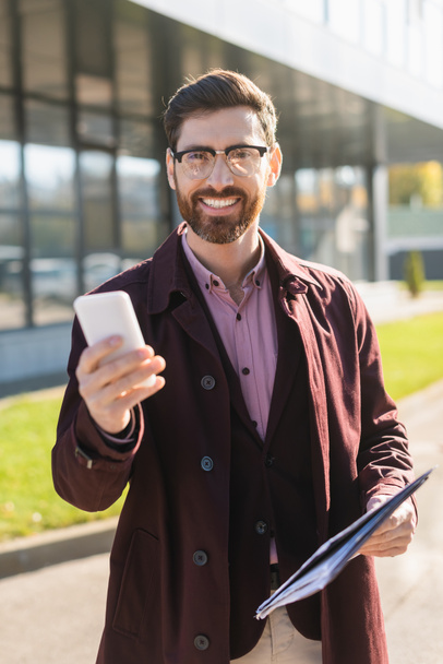 Smiling businessman in trench coat holding smartphone and paper folder outdoors  - Fotoğraf, Görsel