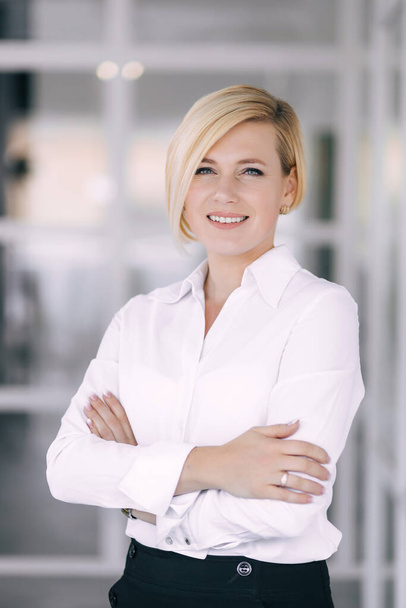 Cheerful successful businessman posing against the background of the office. Proud young woman in an office suit standing with crossed arms and smiling at the camera. Business success concept - Φωτογραφία, εικόνα