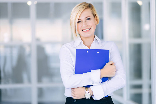 Πορτρέτο μιας νεαρής χαρούμενης επιχειρηματία με λευκό πουκάμισο και φάκελο στα χέρια της. - Φωτογραφία, εικόνα