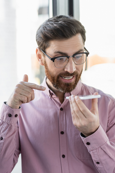 Uomo d'affari in occhiali che registra il messaggio vocale su smartphone e punta con il dito in ufficio  - Foto, immagini