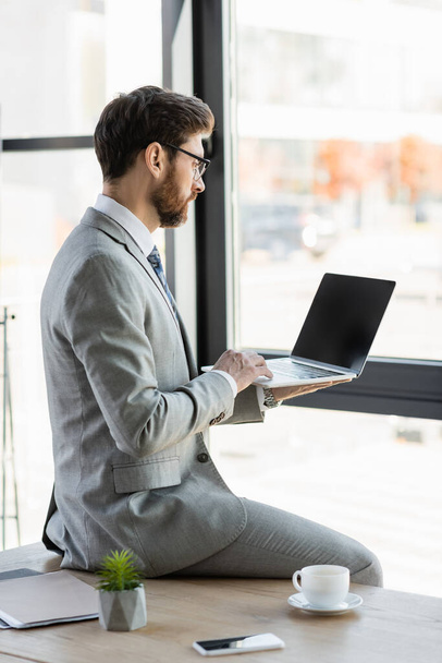 Вид сбоку на бизнесмена, использующего ноутбук с Blank экраном рядом с кофе на столе в офисе  - Фото, изображение