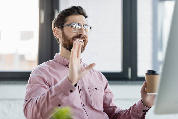 Usmívající se podnikatel držící papírový kelímek během videohovoru na počítači v kanceláři  - Fotografie, Obrázek