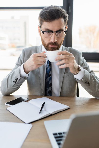 Homme d'affaires tenant tasse et regardant ordinateur portable près de portable dans le bureau  - Photo, image
