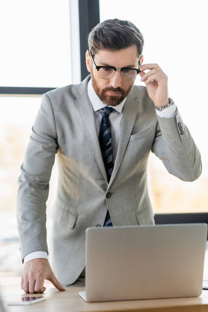 Бизнесмен в очках смотрит на размытый ноутбук в офисе  - Фото, изображение