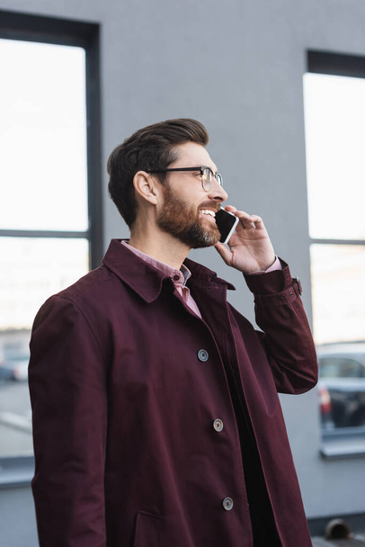 Homme d'affaires joyeux en lunettes et trench coat parler sur smartphone à l'extérieur  - Photo, image