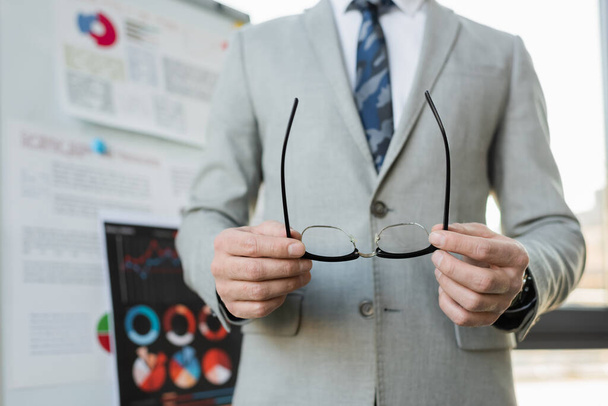Cropped view of businessman holding eyeglasses near blurred flip chart in office  - Photo, Image