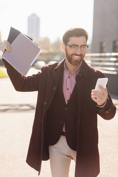 Happy businessman holding paper folders and using smartphone on urban street  - Photo, Image