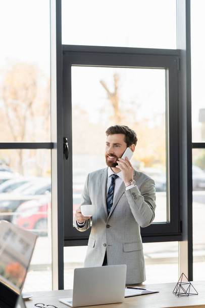 Positive businessman talking on smartphone and holding coffee cup in office  - Foto, afbeelding