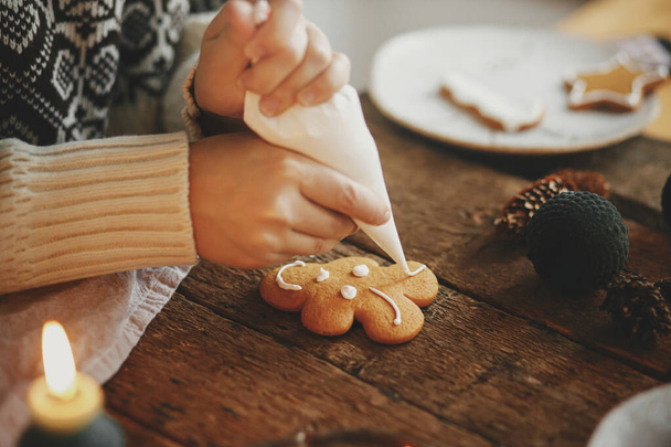 Χέρια διακόσμηση χριστουγεννιάτικο μπισκότο τάρανδο με γλάσο σε ρουστίκ τραπέζι με κερί, μπαχαρικά, διακοσμήσεις. Ατμοσφαιρική κυκλοθυμική εικόνα. Γυναίκα κάνει παραδοσιακά μπισκότα χριστουγεννιάτικο μελόψωμο - Φωτογραφία, εικόνα
