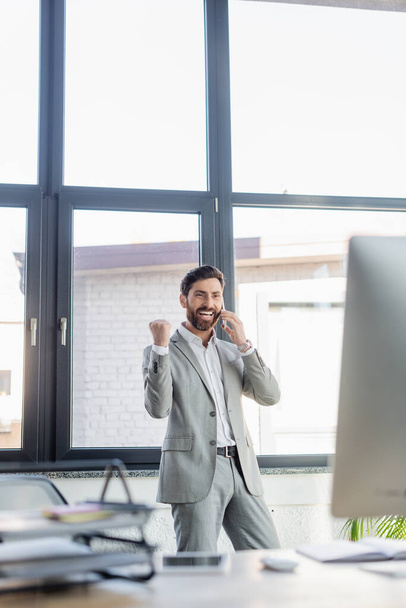 Excited businessman in suit showing yes gesture while talking on smartphone near computer in office  - Foto, Imagem