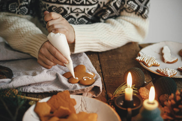 Handen versieren kerst peperkoek man koekje met glazuur op rustieke tafel met servet, specerijen, kaars, decoraties. Sfeervolle humeurige tijd. Traditionele kerstkoekjes bakken met peperkoek - Foto, afbeelding