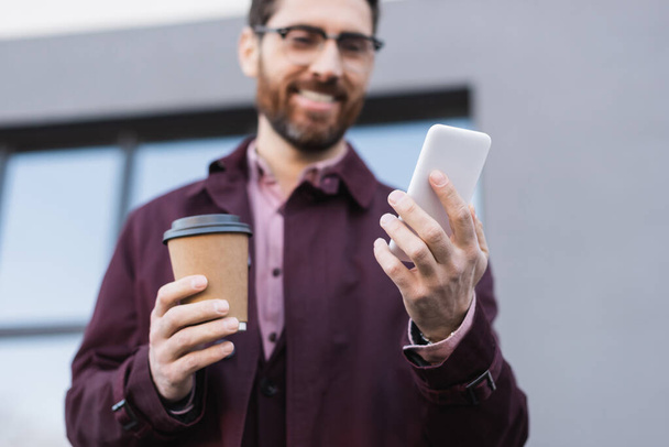 Cellphone in hand of blurred businessman with paper cup outdoors  - Photo, Image