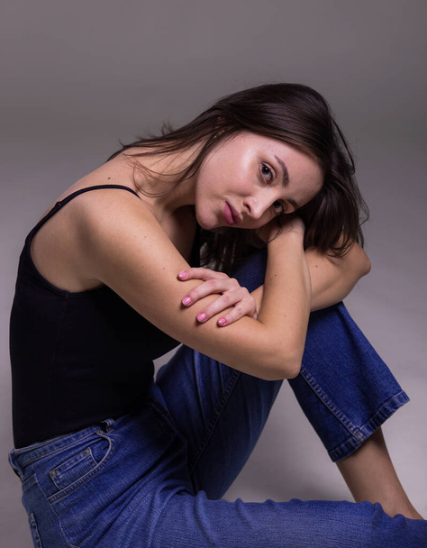 Fashion model sitting on a floor in a jeans and black undershirt barefoot on studio gray background - Photo, Image