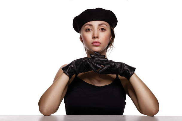 Strategie en oplossingsconcept. Portret van prachtige jonge mode model in zwart baret en lederen handschoenen leunend op de tafel op een witte achtergrond. - Foto, afbeelding