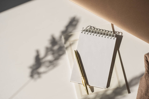 Spiral-Flip-Notizbuch mit leerem Kopierraum, Stift, Laptop in Blumen Sonnenlicht Schatten auf weißem Tisch. Ästhetisch unkonventioneller minimalistischer Arbeitsplatz. Vorlage Künstler, Schriftsteller - Foto, Bild