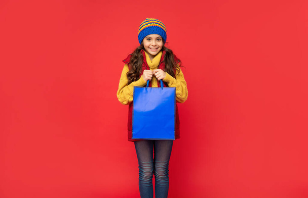 cheerful kid in winter hat hold shopping bags on red background, christmas shopping - Foto, Bild