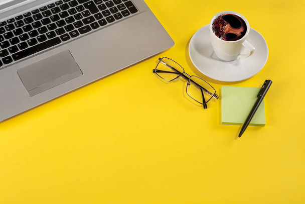 Studio photo avec un fond jaune d'un ordinateur portable à côté d'une tasse de café, verres et stationnaire - Photo, image