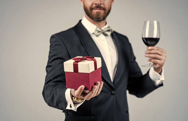 Elegant man with unshaven face cropped view in holiday festive attire hold red wine glass and gift box grey background, celebration - Foto, immagini
