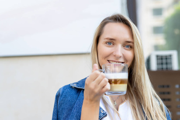 Ritratto di giovane donna felice attraente adulto godere sorridente e tenere tazza di bere caffè cappuccino caldo contro parete caffè di strada in mattina all'aperto. Bella persona femminile wera giacca jeans al di fuori - Foto, immagini