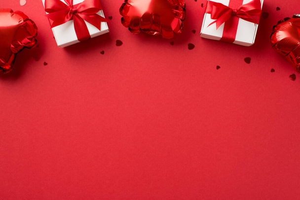 Sevgililer Günü süslemelerinin en üst görüntüsü kırmızı kurdeleli beyaz hediye kutuları kalp şeklinde balonlar ve izole edilmiş kırmızı zemin üzerinde parlak konfetiler. - Fotoğraf, Görsel