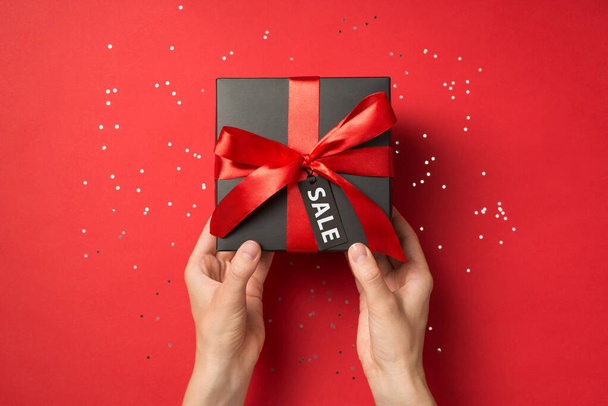 First person top view photo of hands holding black giftbox with red ribbon bow and tag shiny sequins on isolated red background with text on pricetag - Foto, immagini