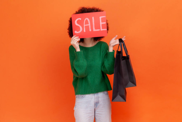 Portrait of unknown woman wearing green casual style sweater standing holding shopping bags, covering face with card with sale inscription. Indoor studio shot isolated on orange background. - 写真・画像