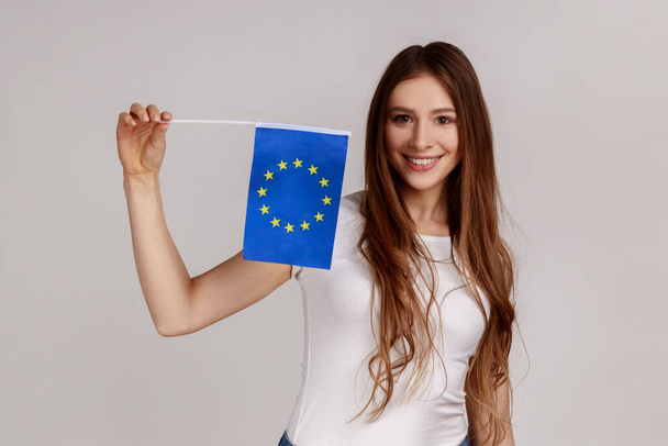 Patriotic woman with dark hair holding eu flag in hand looking at camera with toothy smile, patriotism, flag day celebration, wearing white T-shirt. Indoor studio shot isolated on gray background. - Photo, image