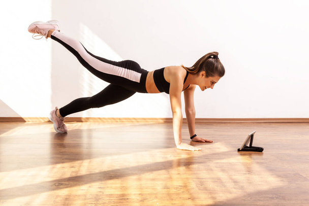 Female doing plank with raised leg, watching tutorial video on tablet, repeat after fitness blogger, wearing black sports top and tights. Full length studio shot illuminated by sunlight from window. - Foto, Imagen