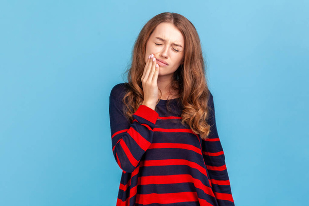 Ill woman wearing striped casual style sweater, touching cheek, closing eyes with expression of terrible suffer from toothache, cavities. Indoor studio shot isolated on blue background. - Foto, immagini