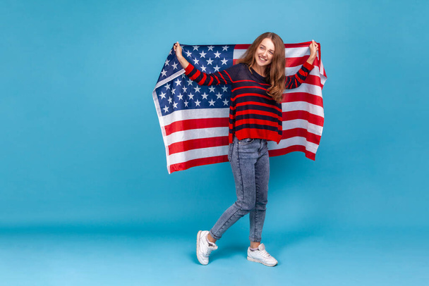 Full length portrait of happy woman in striped casual style sweater, holding USA flag and looking at camera with smile., celebrating national holidays. Indoor studio shot isolated on blue background. - Zdjęcie, obraz