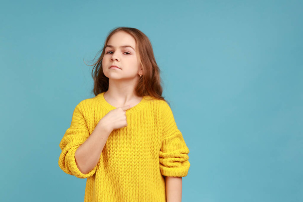 Portrait of little girl pointing herself and looking proudly, boasting of victory, feeling famous, wearing yellow casual style sweater. Indoor studio shot isolated on blue background. - Zdjęcie, obraz