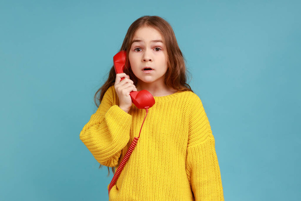 Portrait of little girl holds phone handset surprised by conversation, shocked by unbelievable news, wearing yellow casual style sweater. Indoor studio shot isolated on blue background. - Photo, Image