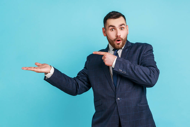 Wow, product advertisement! Portrait of amazed man in suit presenting copy space on palm, showing empty place for commercial text or goods. Indoor studio shot isolated on blue background. - Photo, Image