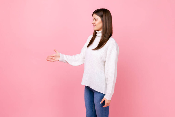 Side view portrait of friendly female standing, giving her hand for handshake, greeting with smile, wearing white casual style sweater. Indoor studio shot isolated on pink background. - Фото, изображение