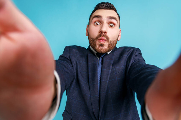 Funny bearded businessman wearing official style suit, taking selfie, looking at camera POV, point of view of photo, sending air kiss. Indoor studio shot isolated on blue background. - Foto, Bild
