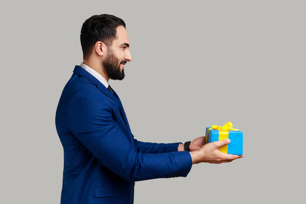 Side view portrait of smiling positive man giving blue wrapped present box, congratulating with holidays, holding gift, wearing official style suit. Indoor studio shot isolated on gray background. - Foto, imagen