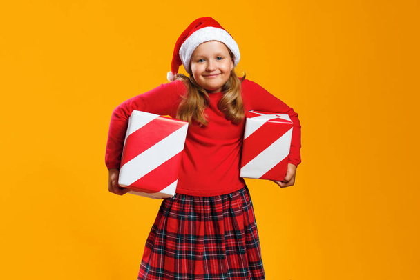 Happy child holding two boxes with Christmas gifts. Little girl in Santa hat on isolated yellow background. - Zdjęcie, obraz