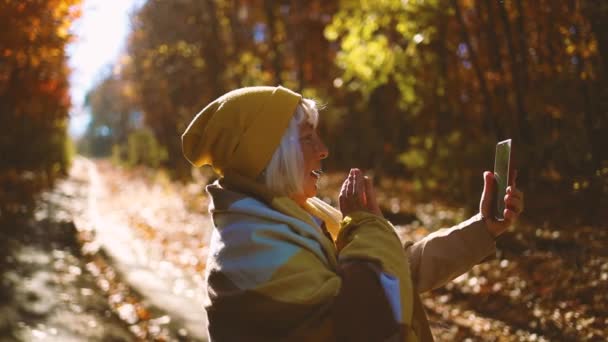 Běloška se cítí šťastný použití smartphone video hovor a pozdravit na sociálních sítích venku v podzimním parku - Záběry, video