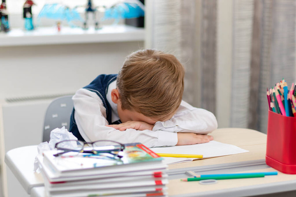 Un lindo chico de primer grado con uniforme escolar en casa durante una pandemia se durmió haciendo deberes en un escritorio con libros y lápices. Enfoque selectivo. Primer plano. Retrato - Foto, imagen