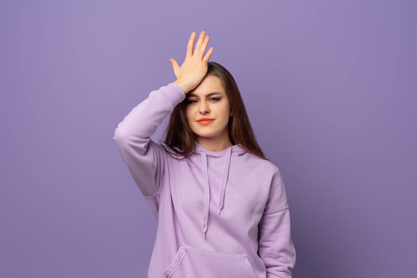 Upset young beautiful woman made a mistake, keeping hand on head, standing over purple background. Young emotional woman in purple hoodie - Fotografie, Obrázek